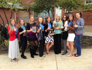 Group of women standing with their thumbs up in front of a red brick building.