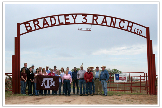 Group under the Bradley 3 Ranch gate sign. They are holding a Texas A&M flag.