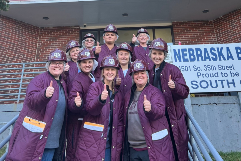 The Fightin' Texas Aggie Meat Judging Team with their maroon lab coats and hard hats on. They are giving a "Gig'em" thumbs up.
