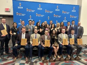 Students sit and stand in front of backdrop holding plaques and banners at a judging competition.