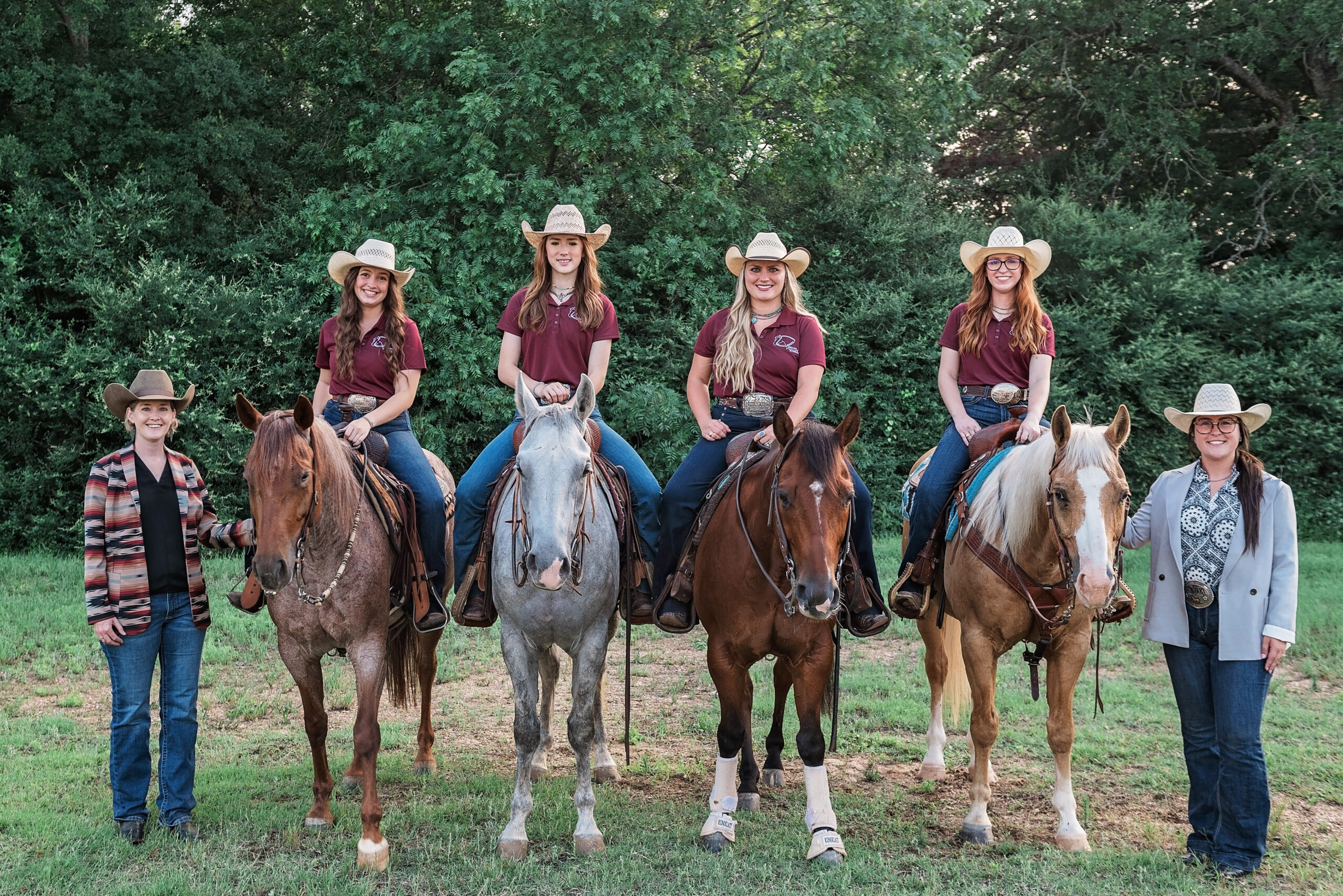 Four woman on horses, and to standing next to them all wearing casual cowboy attire including hats in a natural setting. 