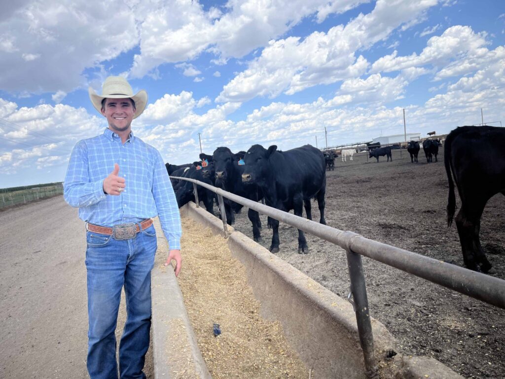 A young man in a cowboy hat, Zach Hoelscher, stands beside a feed bunk in a cattle feedlot with his thumb up during his internship.