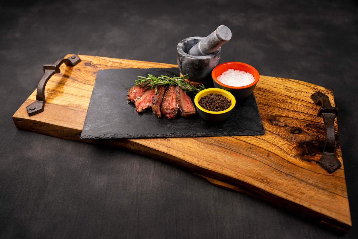 Sliced steak, a sprig of rosemary, and small bowls of peppercorns and salt crystals arranged on a rustic cutting board