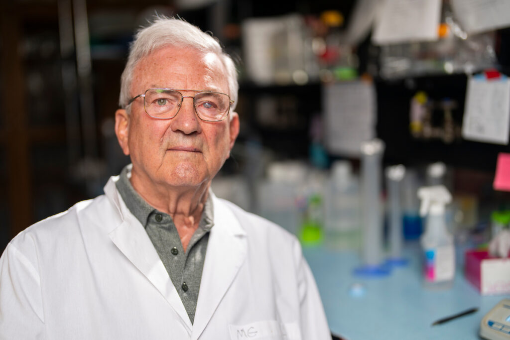 a man in a white lab coat sits with laboratory equipment behind him