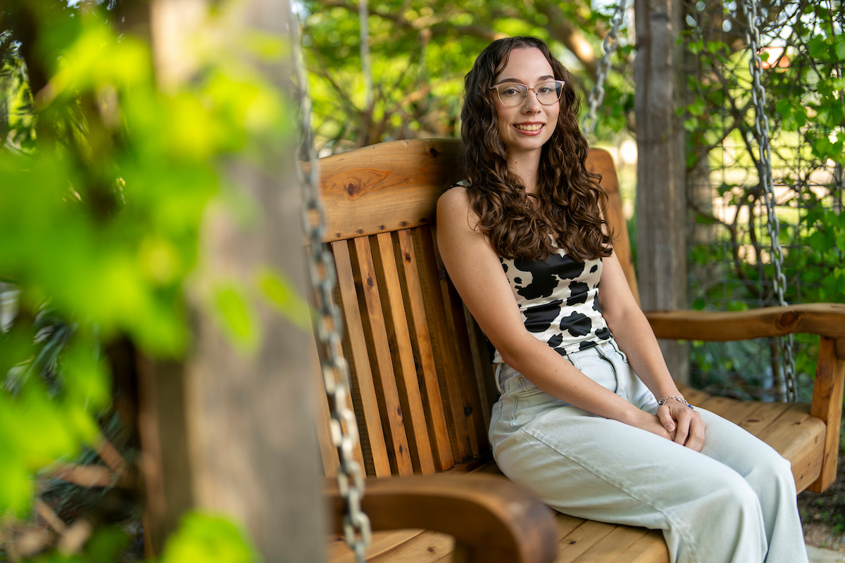 A woman in jeans and a cow print shirt sits on a bench with greenery surrounding her
