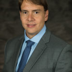 Dr. Reinaldo Cooke in a light blue shirt and a dark grey jacket. He is wearing a dark blue tie.