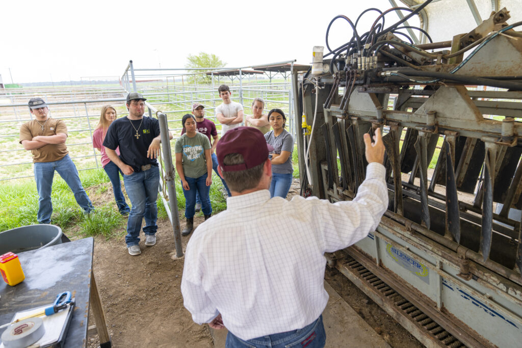 A group of people stand listening to a man in a white shirt and red cap who is explaining equipment in an outdoor, farm-like setting.