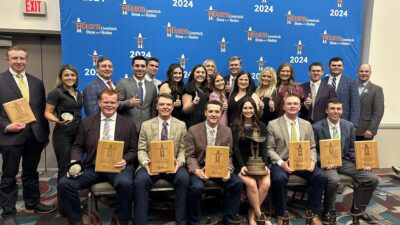 Students sit and stand in front of backdrop holding plaques and banners at a judging competition.