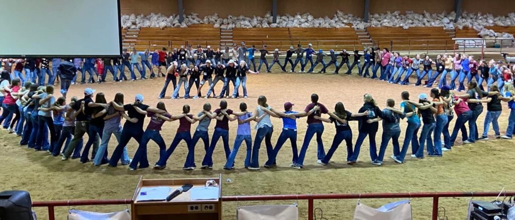 Campers for livestock judging stand in a large circle in an arena, with the camp leaders in the middle giving a presentation.