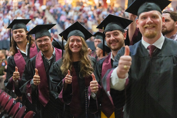 Closeup of 5 Texas A&M students at Texas A&M graduation ceremony, giving a thumbs up which represents the traditional Aggie gig 'em gesture.